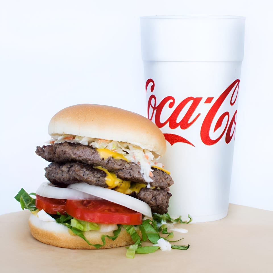 A Morehead Burger Company double cheeseburger with coleslaw, lettuce, tomato, and onion, next to a Coca-Cola cup.